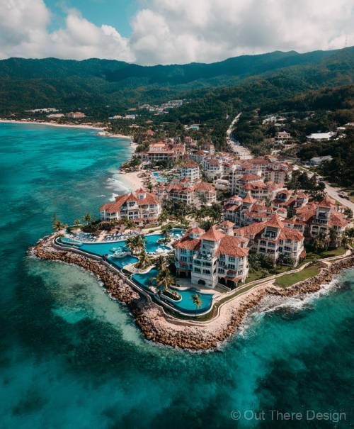 Aerial view of a coastal resort with pools, palm trees, and mountains in the background, embracing turquoise waters.